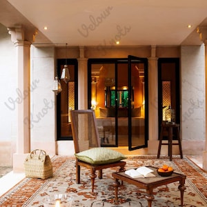 May include: A wooden chair with a cane back and a light green patterned cushion, a low wooden coffee table with an open book and a bowl of oranges, a woven basket, and a small wooden stool on a large patterned rug under a covered patio.