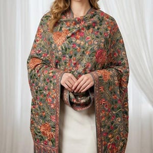 Puede incluir: Un chal gris con un intrincado bordado floral multicolor en tonos naranja, rosa, verde y amarillo. La prenda rectangular muestra un patrón denso de flores en flor y enredaderas a lo largo de toda la superficie, con un borde de color gris sólido.