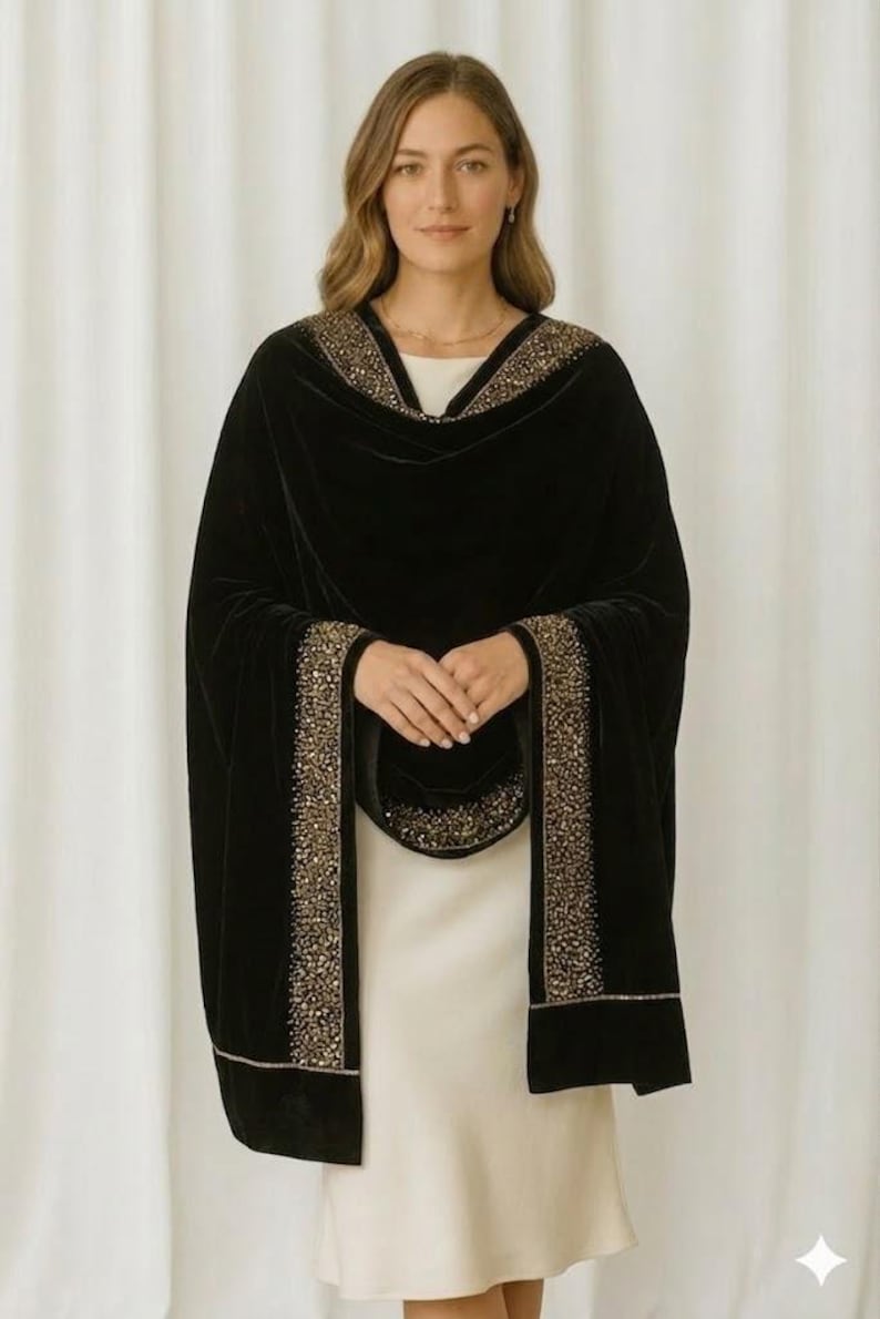 Puede incluir: Un chal de terciopelo negro con ribete de cuentas doradas. El chal se lleva sobre un vestido blanco. Las cuentas doradas se concentran en los bordes y el escote. Un accesorio lujoso.