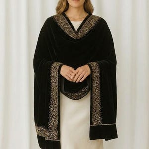 Puede incluir: Un chal de terciopelo negro con adornos dorados. El chal est&aacute; drapeado sobre un vestido blanco. Los adornos dorados est&aacute;n a lo largo de los bordes y el escote del chal. El chal tiene mangas largas.