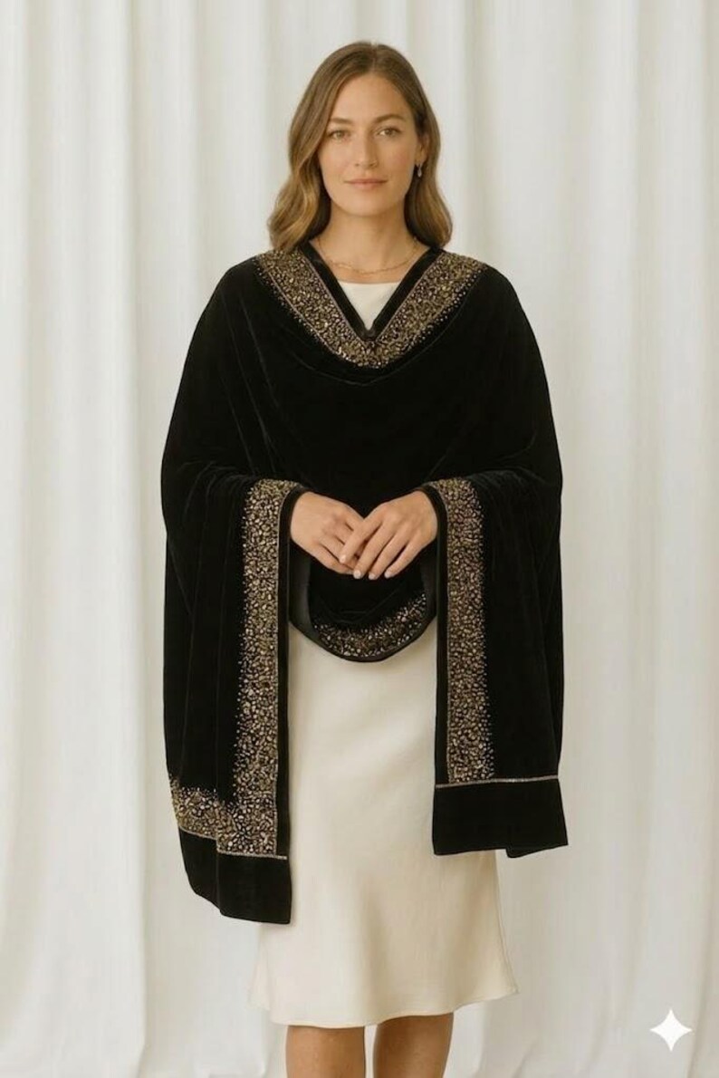 Puede incluir: Un chal de terciopelo negro con adornos dorados. El chal est&aacute; drapeado sobre un vestido blanco. Los adornos dorados est&aacute;n a lo largo de los bordes y el escote del chal. El chal tiene mangas largas.