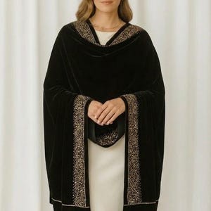 Puede incluir: Un chal de terciopelo negro con ribete de cuentas doradas. El chal est&aacute; drapeado sobre un vestido blanco. El ribete recorre los bordes del chal y alrededor del escote. Un accesorio lujoso.