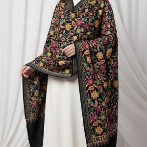 May include: A black shawl featuring an intricate, multi-colored floral embroidery pattern in shades of yellow, pink, and green. The rectangular wrap has a dense, detailed botanical design covering the fabric and a decorative border along the edges.