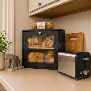 May include: A black bread box with a glass door, filled with loaves of bread, sits on a countertop next to a stainless steel toaster. A wooden cutting board and a small elephant statue are also visible.
