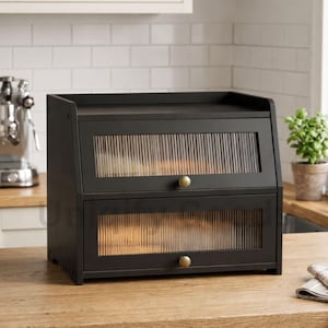 May include: A black wooden bread box with two compartments and ribbed glass doors. The bread box has brass knobs and sits on a wooden countertop. The background includes a white tiled wall and a small potted plant.