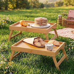 May include: Two bamboo breakfast trays with folding legs are set on a grassy lawn. One tray holds pancakes and orange slices, the other has croissants and a cup of coffee. A picnic basket and blanket are in the background.