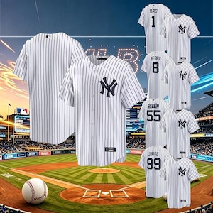 Op de afbeelding: Witte honkbalshirts met krijtstrepen en het logo van de New York Yankees. Shirts tonen namen en nummers van spelers, waaronder "DAD 1", "BERRA 8", "RODON 55" en "JUDGE 99". Een honkbal ligt op het veld.