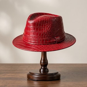 Könnte beinhalten: Ein leuchtend roter Fedora-Hut mit einer strukturierten Krokodilleder-Optik auf der gesamten Oberfläche. Der Hut verfügt über ein passendes rotes Band um die Basis der Krone, eine strukturierte Krempe und ein klassisches eingedrücktes Design.