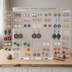 Könnte beinhalten: Eine Präsentation verschiedener gehäkelter Ohrringe in unterschiedlichen Formen und Farben, darunter Herzen, Blätter und geometrische Designs. Der Display enthält den Text "100 ULTIMATE CROCHET EARRING PATTERNS - COMPLETE DIGITAL COLLECTION". Ein Knäuel Garn und eine Häkelnadel sind ebenfalls vorhanden.