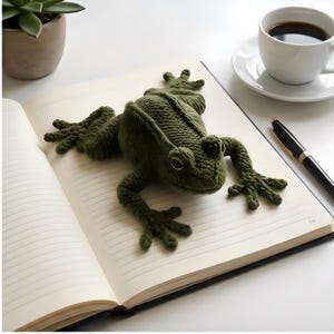May include: An olive green crochet frog sits on an open notebook. The frog is detailed with bulging eyes and textured skin. A cup of coffee and a pen are also on the white surface, suggesting a workspace or desk setting.