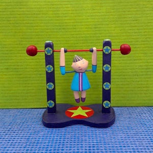 May include: A whimsical figurine of a character doing a pull-up on a red bar. The figure is wearing a blue dress with purple and white stripes. The base and support beams are dark blue with star accents and red spheres.