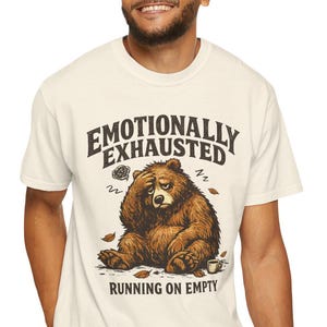 Pode incluir: Uma t-shirt de manga curta em tom off-white com um gráfico de um urso castanho sentado com a mão na testa. O texto "EMOTIONALLY EXHAUSTED" aparece acima do urso e "RUNNING ON EMPTY" está impresso abaixo da ilustração do urso.