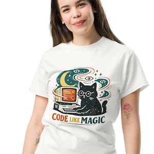 Może przedstawiać: Biały t-shirt z grafiką przedstawiającą czarnego kota w okularach siedzącego przy komputerze w stylu vintage. Wzór zawiera półksiężyc, gwiazdy i symbole kodu w kolorach morskim, pomarańczowym i czarnym. Pod kotem znajduje się napis CODE LIKE MAGIC.