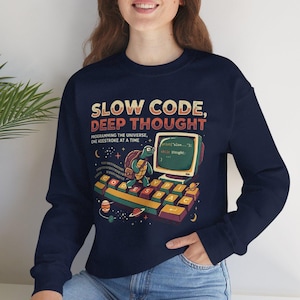 May include: A navy blue long-sleeve sweatshirt featuring a graphic print with the text "SLOW CODE, DEEP THOUGHT" and "PROGRAMMING THE UNIVERSE, ONE KEESTROKE AT A TIME". The design includes a turtle on a keyboard, a computer monitor, and planets.