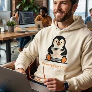 Peut inclure: Un sweat à capuche couleur crème arborant un motif de pingouin de dessin animé portant des lunettes et travaillant sur un ordinateur portable, avec le texte "Powered by Linux" imprimé en dessous. Le sweat a des manches longues et une poche.