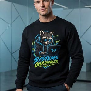 May include: A black long-sleeve sweatshirt featuring a graphic of a raccoon wearing blue sunglasses and headphones while using a laptop. The text "SYSTEMS OVERTHINKER" is printed in blue and yellow below the raccoon illustration.