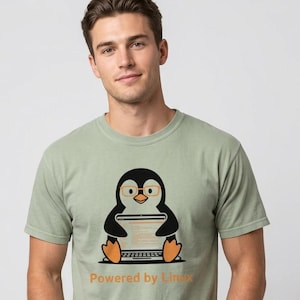 May include: Sage green t-shirt featuring a cartoon penguin wearing glasses and using a laptop. The text "Powered by Linux" is printed in orange below the penguin. The shirt is worn by a person.
