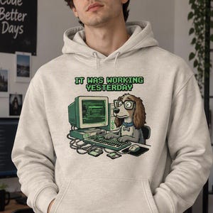 Può includere: Una felpa con cappuccio beige chiaro con un'illustrazione grafica di un cane marrone con gli occhiali, seduto davanti a un computer vintage con il testo IT WAS WORKING YESTERDAY stampato in verde sopra il cane e il computer.