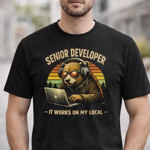May include: A black t-shirt featuring a graphic of a brown bear wearing headphones and sunglasses while typing on a laptop. The text reads SENIOR DEVELOPER and IT WORKS ON MY LOCAL, set against a circular sunset background with silhouettes of trees.