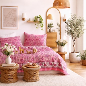 May include: A bed featuring a pink quilt and two matching pillow shams with a floral pattern. The bed is positioned in a room with two round woven side tables, a large potted olive tree, a tall arched mirror, and a framed line art print.