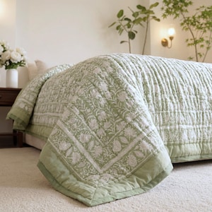 May include: A light green and white floral patterned quilt on a bed. The quilt has a detailed design with a solid light green border. The bed is in a well-lit room with plants and a lamp.