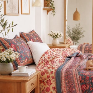 May include: A quilted bedspread and two matching pillow shams featuring a floral pattern in shades of red, blue, and cream. The bedding set is displayed on a bed with a wooden nightstand holding a white ceramic vase of flowers and a book.