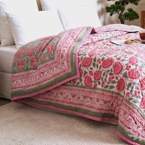 Può includere: Una trapunta floreale con un design rosa e verde. La trapunta presenta grandi fiori rosa su sfondo bianco, incorniciati da un bordo verde e finiture rosa. La biancheria da letto è su un letto bianco con cuscini bianchi.
