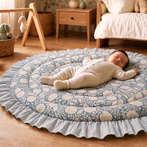 May include: A round, quilted play mat featuring a blue and beige floral pattern with a ruffled edge in a blue and white pinstripe fabric. A baby wearing a cream-coloured knitted onesie is lying on the mat, which is placed on a wooden floor.