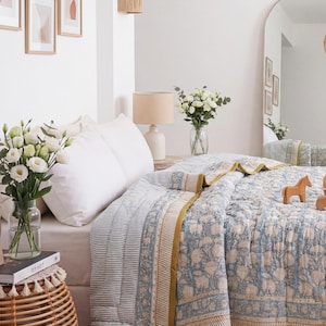 May include: A quilted bedspread featuring a light blue and white floral block print pattern with mustard yellow border accents. The bedding is draped over a bed with white pillows, accompanied by wooden animal figurines on the surface.