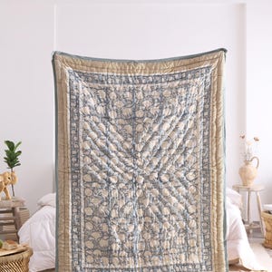 Könnte beinhalten: Eine rechteckige, gesteppte Tagesdecke mit einem sich wiederholenden blauen Blumenmuster auf cremefarbenem Grund. Die Decke ist von einem gestreiften Rand in Beige- und Blautönen umrahmt und weist durchgehend sichtbare Steppnähte auf.