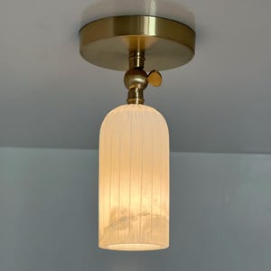 May include: A modern ceiling light fixture with a gold-colored base and a frosted, cylindrical glass shade. The shade has vertical lines and emits a warm, inviting glow. The fixture is mounted on a white ceiling.