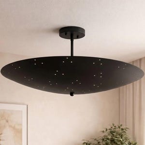 Könnte beinhalten: Eine moderne, schwarze Deckenleuchte. Die Leuchte hat einen kreisförmigen Sockel und eine lange, dünne Stange, die mit einem großen, oval geformten Schirm verbunden ist. Der Schirm ist mit kleinen Löchern perforiert, durch die das Licht scheint.