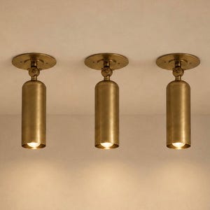Peut inclure: Trois spots de plafond en laiton. Chaque luminaire est composé d'une base circulaire, d'un corps cylindrique et d'une petite source de lumière focalisée. Les lumières sont espacées uniformément et montées sur un plafond de couleur neutre.