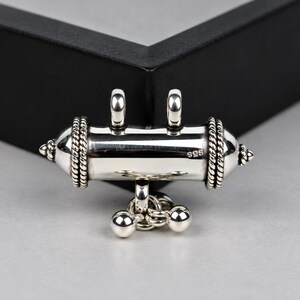 May include: A polished silver cylindrical pendant featuring a smooth central body, two top loops for suspension, and decorative rope-textured bands at each end. The piece includes a small dangling charm with three silver spheres and is stamped 925.
