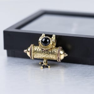 Puede incluir: Un colgante cilíndrico de tono dorado con intrincados patrones de volutas grabados, una piedra cabujón negra central engastada en un marco de cuentas y tres pequeñas cuentas negras colgando del centro inferior.