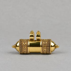 May include: A gold-toned cylindrical pendant featuring a smooth central band flanked by two textured sections with beaded and circular patterns. The piece includes two small vertical loops at the top for attachment and rounded, pointed ends.