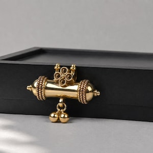 May include: A rectangular black box featuring a centered gold-toned cylindrical pendant. The pendant has a smooth central body, textured rope-like ends, a four-petaled floral top element, and two small dangling spherical charms at the bottom.