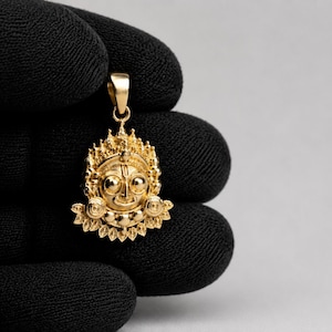 May include: A gold pendant featuring a detailed, stylised face with large circular eyes, a central forehead mark, and an ornate, radiating crown-like border. The piece includes a small, smooth bail at the top for attachment to a chain or cord.