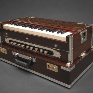 May include: A dark brown harmonium with a keyboard and multiple knobs. The instrument has a rectangular shape with silver metal trim and a carrying handle. The top of the harmonium has carved details.