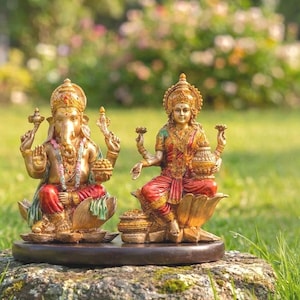 May include: Two detailed statues of Hindu deities, Ganesha and Lakshmi, are displayed outdoors. Ganesha, with an elephant head, wears red and gold clothing. Lakshmi, in red and gold, sits on a lotus. Both figures are on a dark brown base.