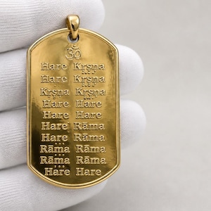 Könnte beinhalten: Ein goldfarbener Anhänger in Form einer Erkennungsmarke mit einem eingravierten Om-Symbol an der Oberseite und dem Hare-Krishna-Mantra in zehn Zeilen. Der rechteckige Anhänger hat abgerundete Ecken, eine polierte Oberfläche und eine Öse.