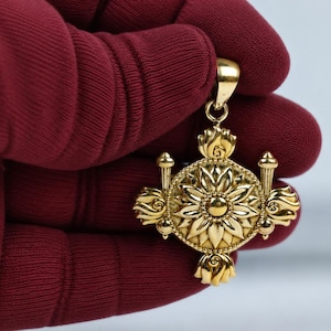 Puede incluir: Un colgante de oro que presenta un medallón circular central con un motivo floral en relieve, rodeado por cuatro elementos decorativos simétricos y una pequeña anilla en la parte superior. La pieza tiene un acabado pulido y metalurgia detallada.