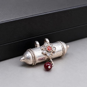 Puede incluir: Un colgante cilíndrico de tono plateado con un adorno central en forma de flor con piedras transparentes y una piedra central de color rojo anaranjado. Una pequeña cuenta redonda de color rojo oscuro cuelga de la parte inferior y dos anillas.