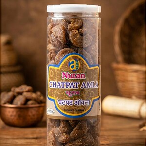 Op de afbeelding: Een doorzichtige plastic pot gevuld met bruin, gedroogd fruit, met het label "Nutan Chatpat Amla". De pot heeft een witte deksel en een goud en blauw etiket met tekst in meerdere talen. Product van India.