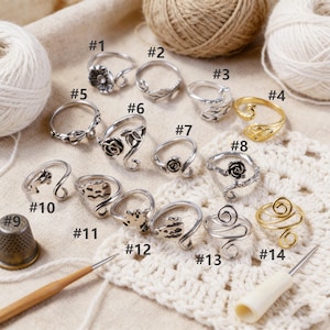 Könnte beinhalten: Eine Sammlung von vierzehn Ringen mit verschiedenen Designs, darunter Blumen-, Blatt- und abstrakte Muster. Die Ringe sind silber- und goldfarben, einige mit aufwendigen Details. Ein Fingerhut und eine Häkelnadel sind ebenfalls vorhanden.