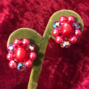 May include: A pair of vintage clip-on earrings. Each earring has a central oval red bead, surrounded by smaller pink beads and iridescent crystals. The earrings are displayed on a green velvet stand against a red velvet background.
