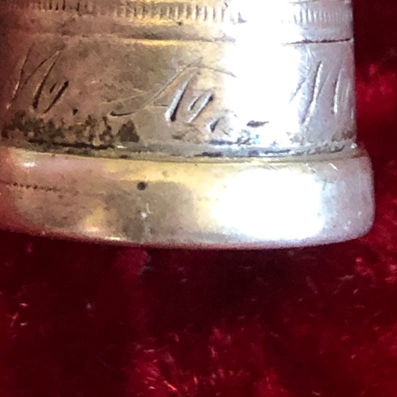 May include: Close-up of a silver thimble with engraved script. The thimble has a slightly tarnished appearance, with visible wear and tear. The script appears to be a signature or initials. The thimble is set against a red background.