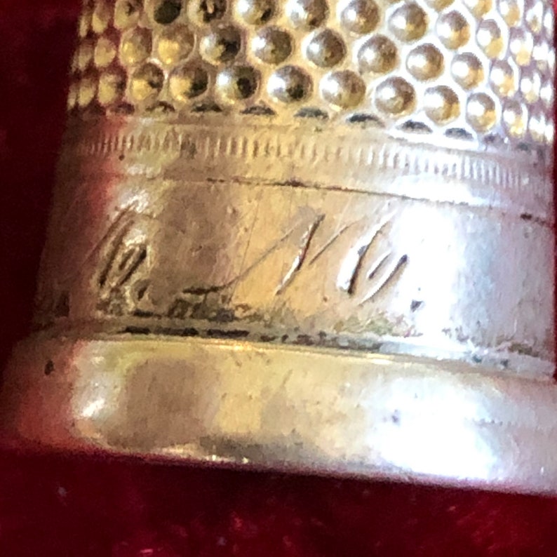 May include: Close-up of a silver thimble with a textured top and engraved markings. The thimble has a polished finish and is set against a red background. The engraved text appears to be initials or a signature.