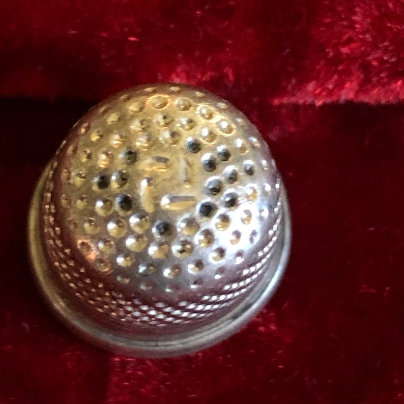 May include: A silver thimble with a rounded top covered in small dimples. The base has a decorative, textured band. The thimble is set against a deep red background, creating a contrast that highlights its metallic sheen. A sewing tool.