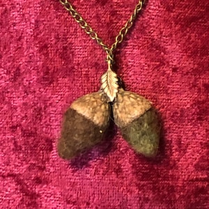 May include: A necklace featuring two acorn pendants with dark brown caps and tan bodies. The acorns are connected by a gold-colored leaf charm and suspended from a delicate chain. The background is a deep red fabric.